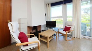 Una sala de estar moderna con chimenea, dos sillones blancos con cojines rojos, una mesa de centro de madera y un televisor de pantalla plana.