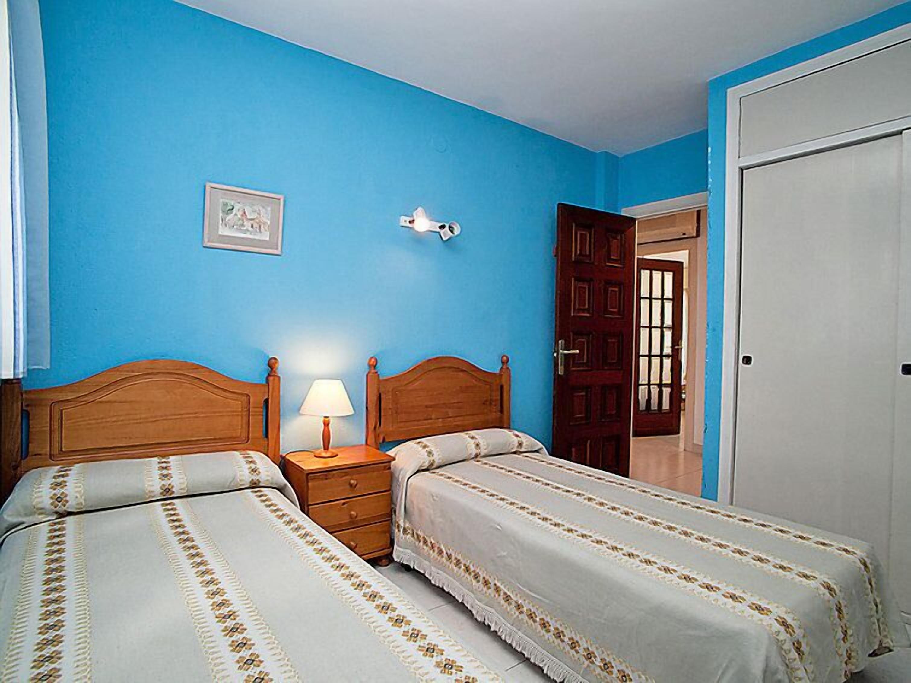 A bedroom with two beds, a nightstand, a lamp, a framed picture on the wall, and a door leading to another room.
