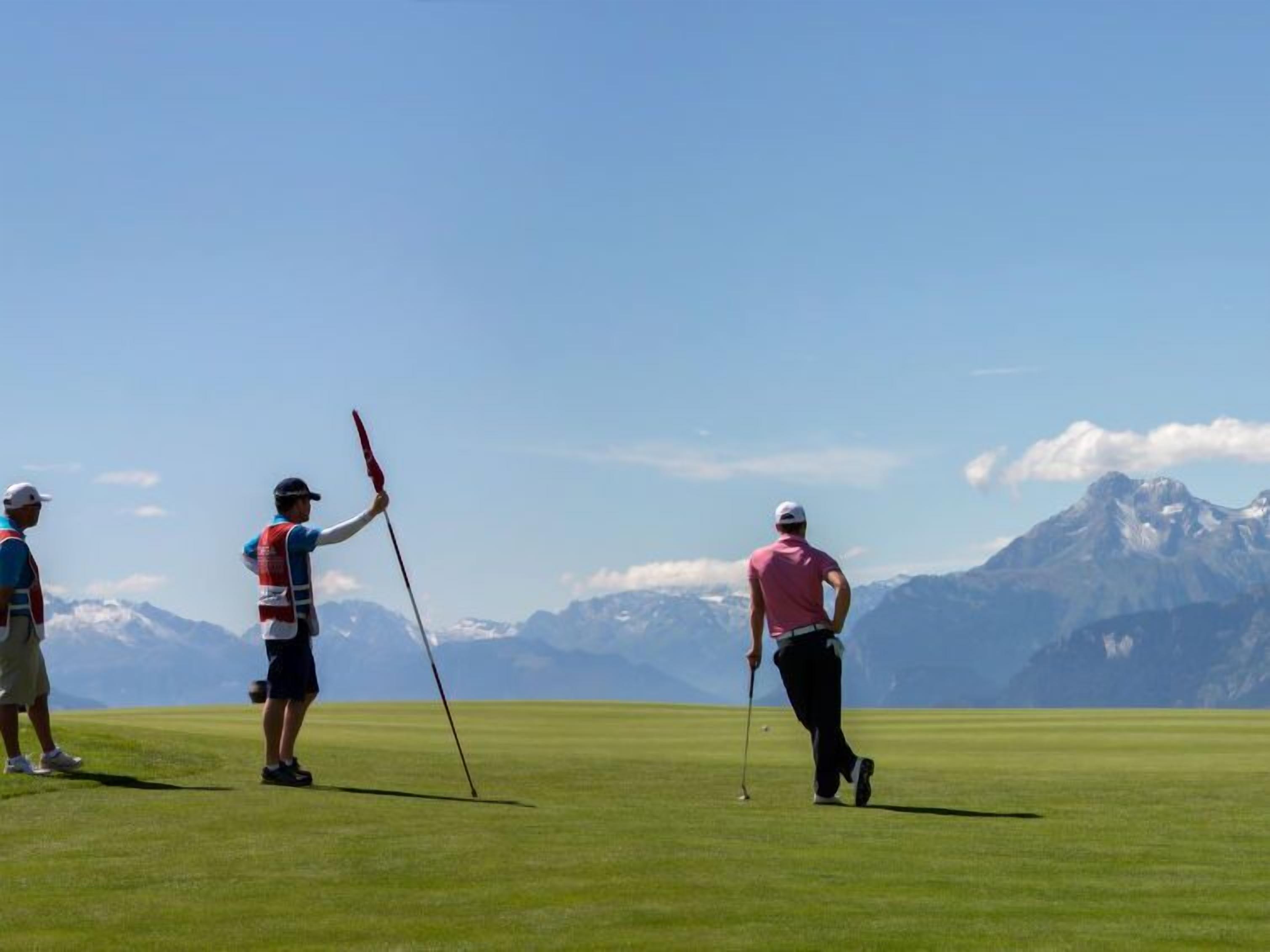 Un campo de golf con jugadores en movimiento, cielo despejado y montañas al fondo.