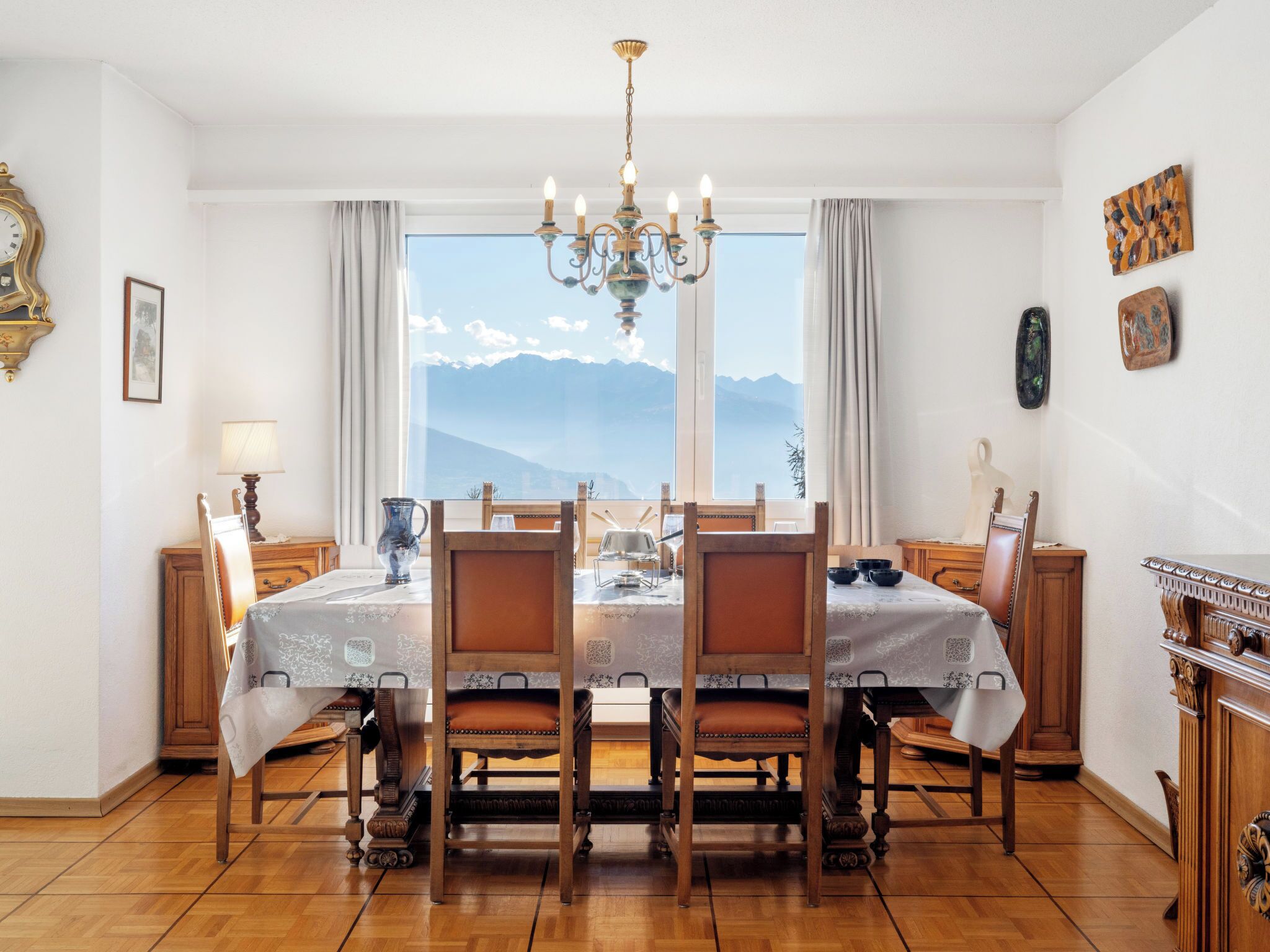 Ein Esszimmer mit Tisch für vier Personen, Holzsesseln, einer Pendelleuchte und Blick auf Berge durch große Fenster.