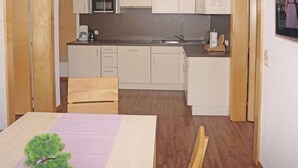 Eine kompakte Küche mit Mikrowelle, Backofen und Spüle. Ein Esstisch mit einem Stuhl und ein kleiner Holztisch sind vorhanden. Der Raum verfügt über Holzboden und helle Wände.