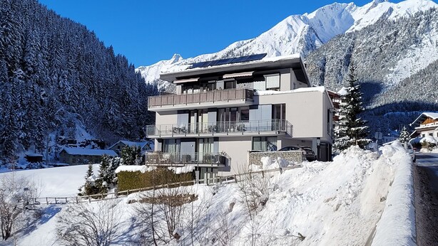 Ein modernes, zweistöckiges Gebäude mit Balkonen, umgeben von schneebedeckten Bergen und Bäumen.