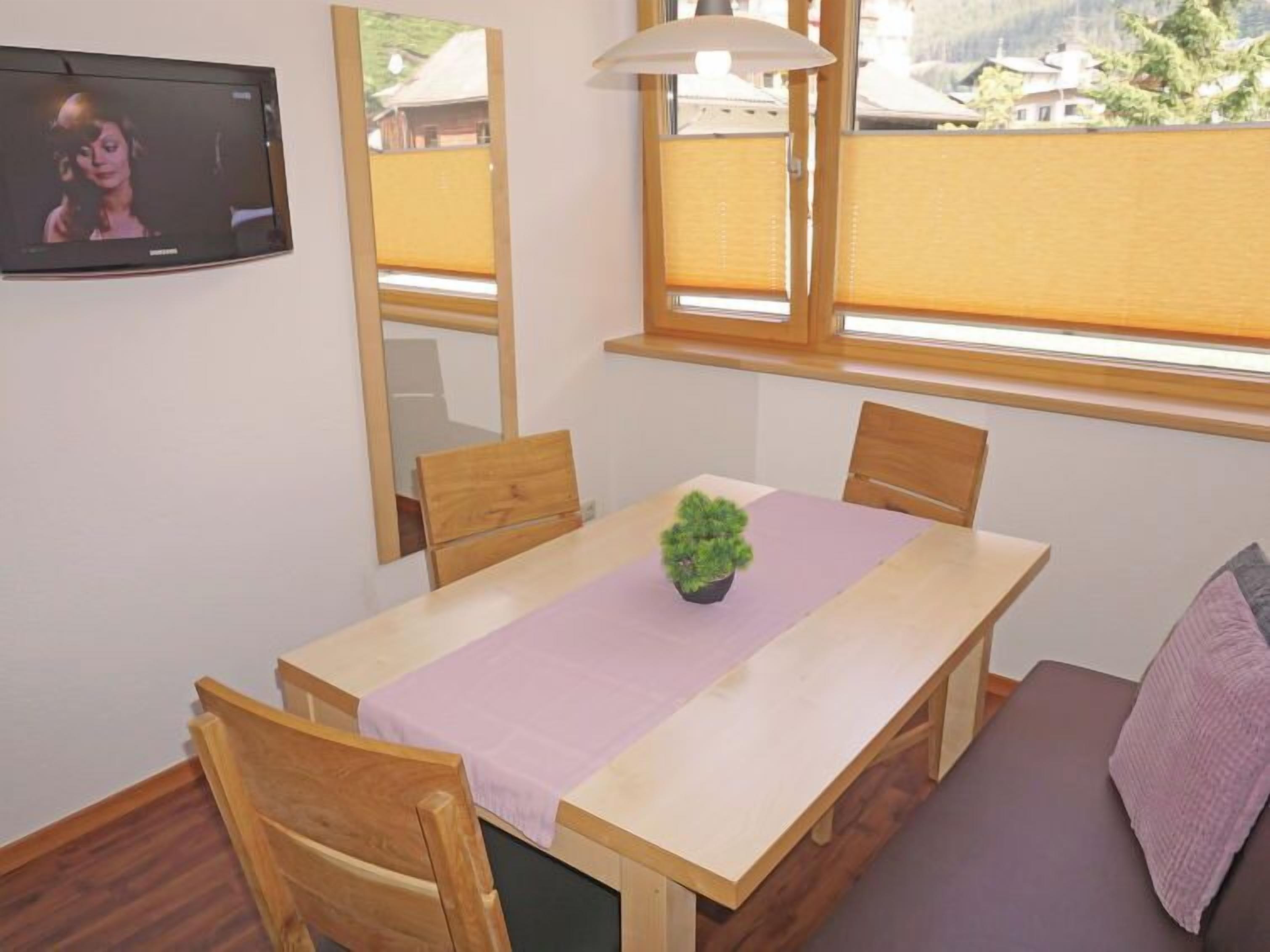 A room with a wooden dining table, chairs, a small potted plant, a sofa, and a TV mounted on the wall.