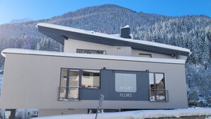 Ein modernes Gebäude mit Flachdach und Balkon, umgeben von schneebedeckten Bäumen und einem Berg im Hintergrund.