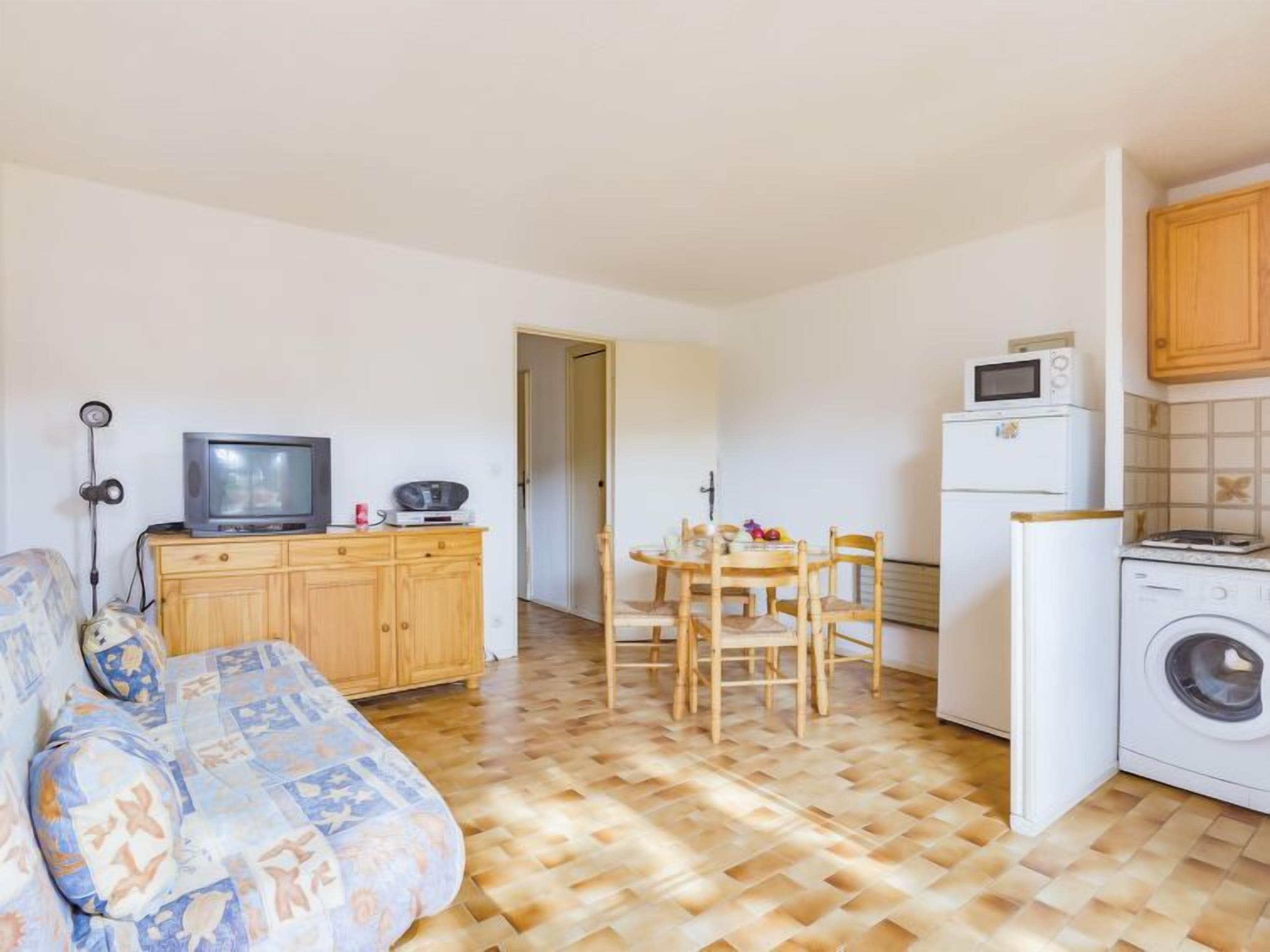 A compact living space with a sofa, a dining table with chairs, a kitchen area including a microwave and washing machine, and a television on a wooden cabinet.