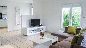 Een moderne woonkamer met een witte tv-kast, een flatscreen televisie, een glazen salontafel en een bruine bank met groene kussens.
