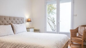 Een slaapkamer met een groot bed, een nachtkastje met een lamp, een rieten stoel en een schuifdeur naar een balkon.
