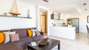 Una sala de estar moderna con sofá, mesa de centro y objetos decorativos. Se ve una cocina al fondo, con una isla central y electrodomésticos.