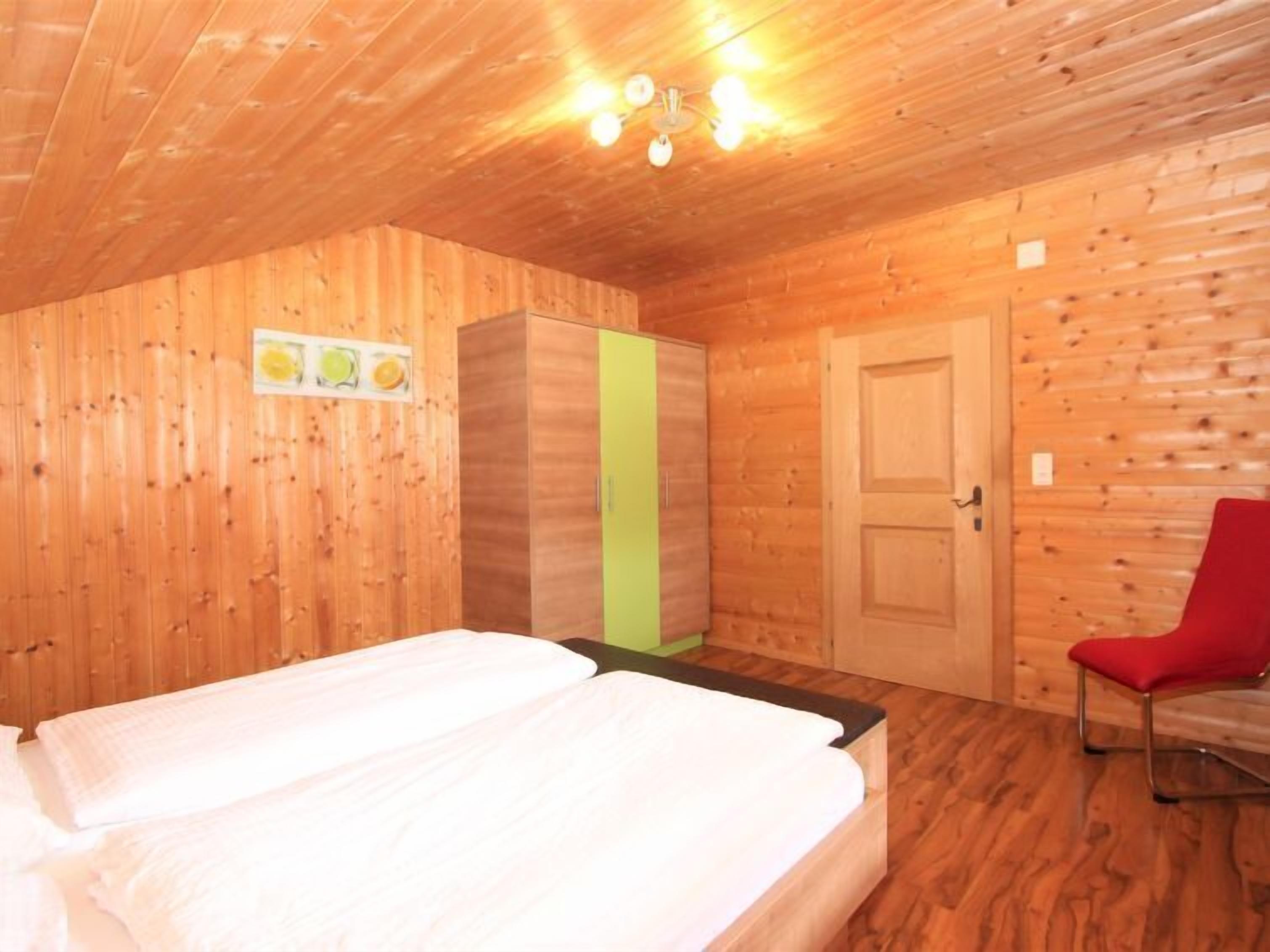 A wooden room with a bed, a green wardrobe, a red chair, and a ceiling light fixture.