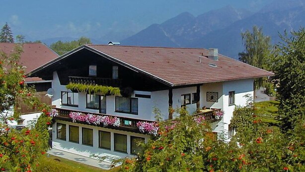 Een huis met twee verdiepingen, een schuin dak, een balkon met bloemen en een berg op de achtergrond.