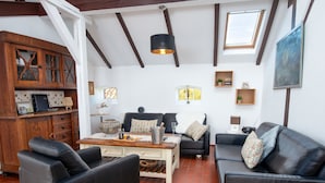 Uma sala de estar moderna com pé direito alto, vigas aparentes e um claraboia. Há dois sofás de couro preto, uma mesa de centro de madeira e uma mesa de jantar com um vaso e velas.