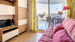 A modern living room with a flat-screen TV, wooden shelving, a pink sofa, and a balcony with yellow curtains.