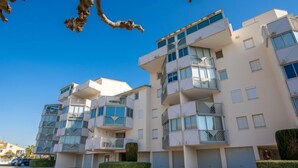 A modern multi-story residential building with balconies, a parking area, and a clear blue sky.