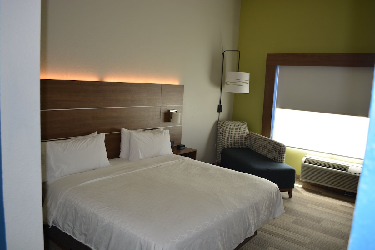 A hotel room with a large bed, a nightstand, a lamp, a chair, and an air conditioning unit.