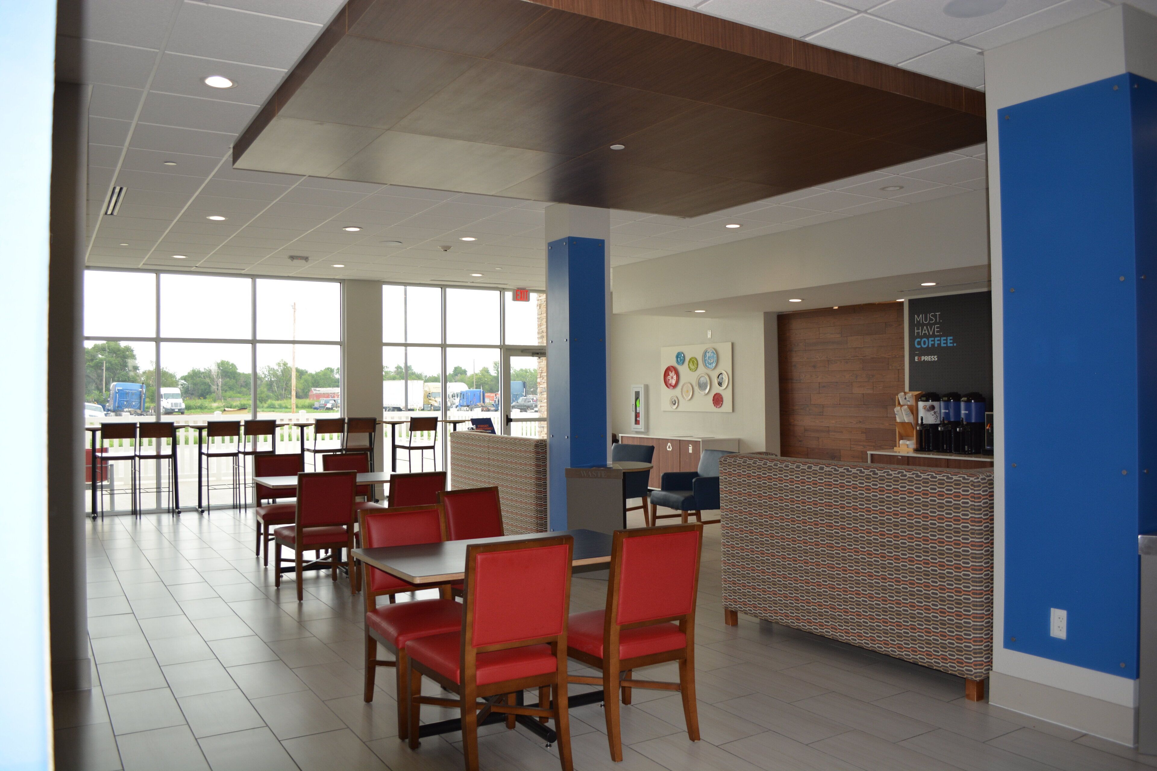 A spacious cafeteria with red chairs and tables, a coffee bar, and large windows offering a view of the parking lot.
