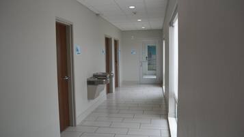 A clean, modern hallway with white tiled floors, wooden doors, and a hand sanitizer dispenser.