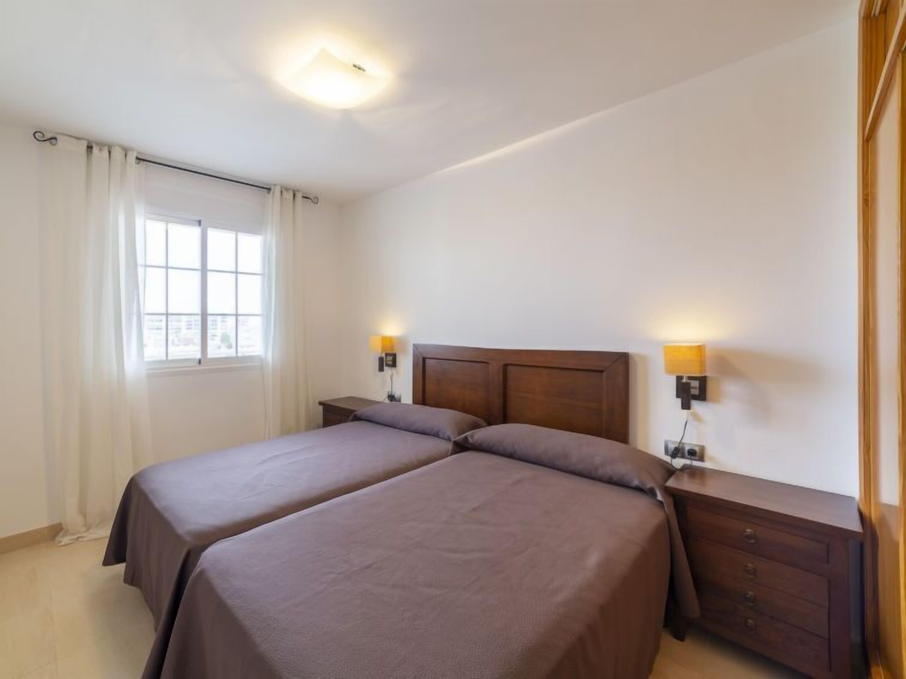 Un lit double avec deux tables de chevet, une fenêtre avec des rideaux et un luminaire fixé au mur.
