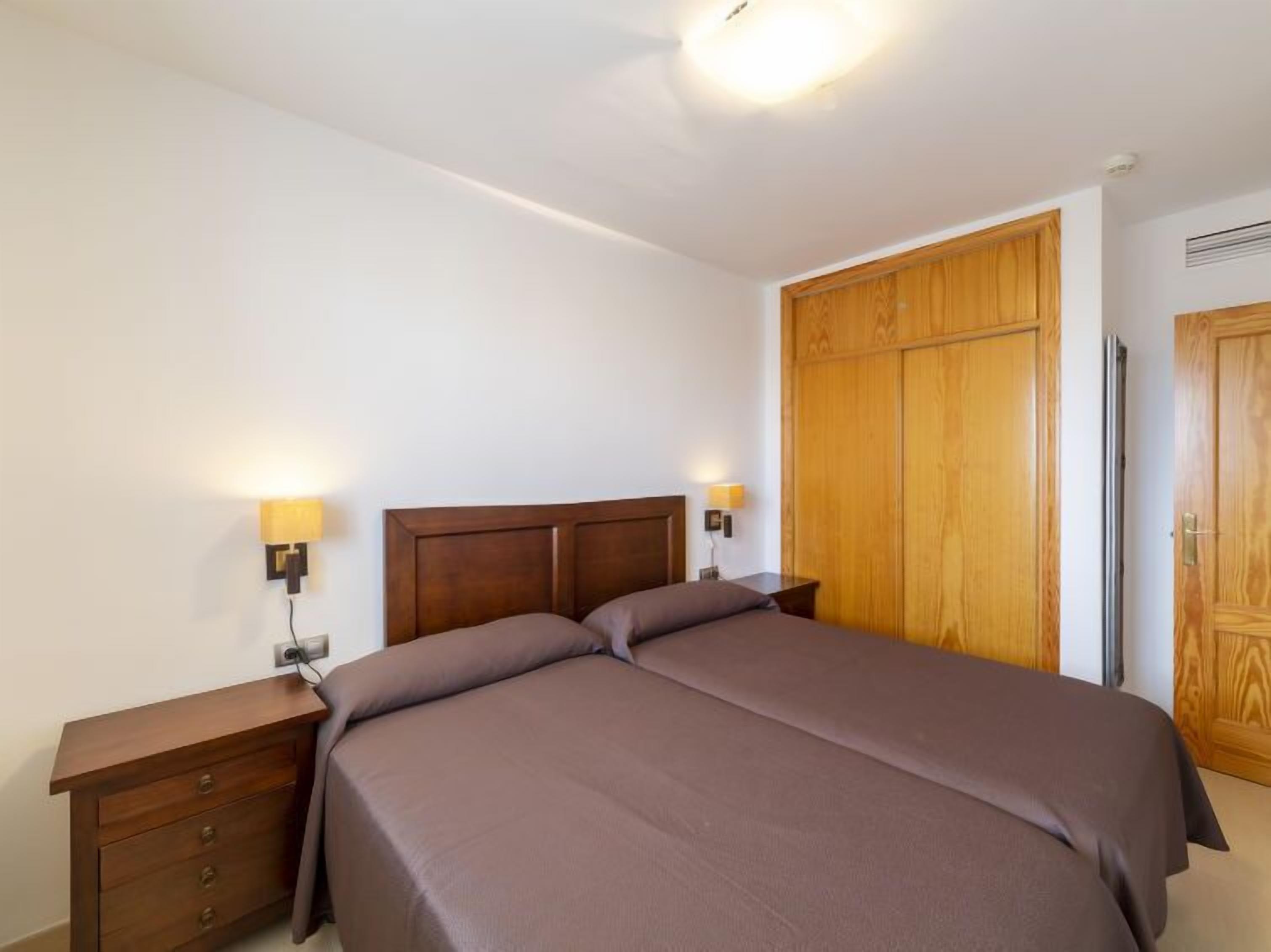 Een slaapkamer met een tweepersoonsbed, een houten garderobe, nachtkastjes en wandlampen.