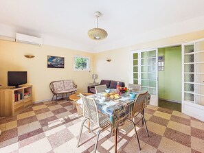 Ein Wohnzimmer mit Fernseher, Bücherregalen, einem Esstisch mit Stühlen, einer Couch und einem gemusterten Boden.