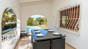A balcony with a table set for two, a window with decorative bars, and a view of a swimming pool and greenery.