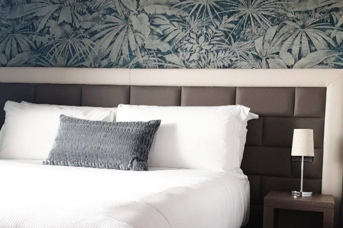 Una cama bien tendida con sábanas blancas y un cojín gris texturizado, una mesita de noche con una lámpara, y una pared con un estampado de hojas tropicales.