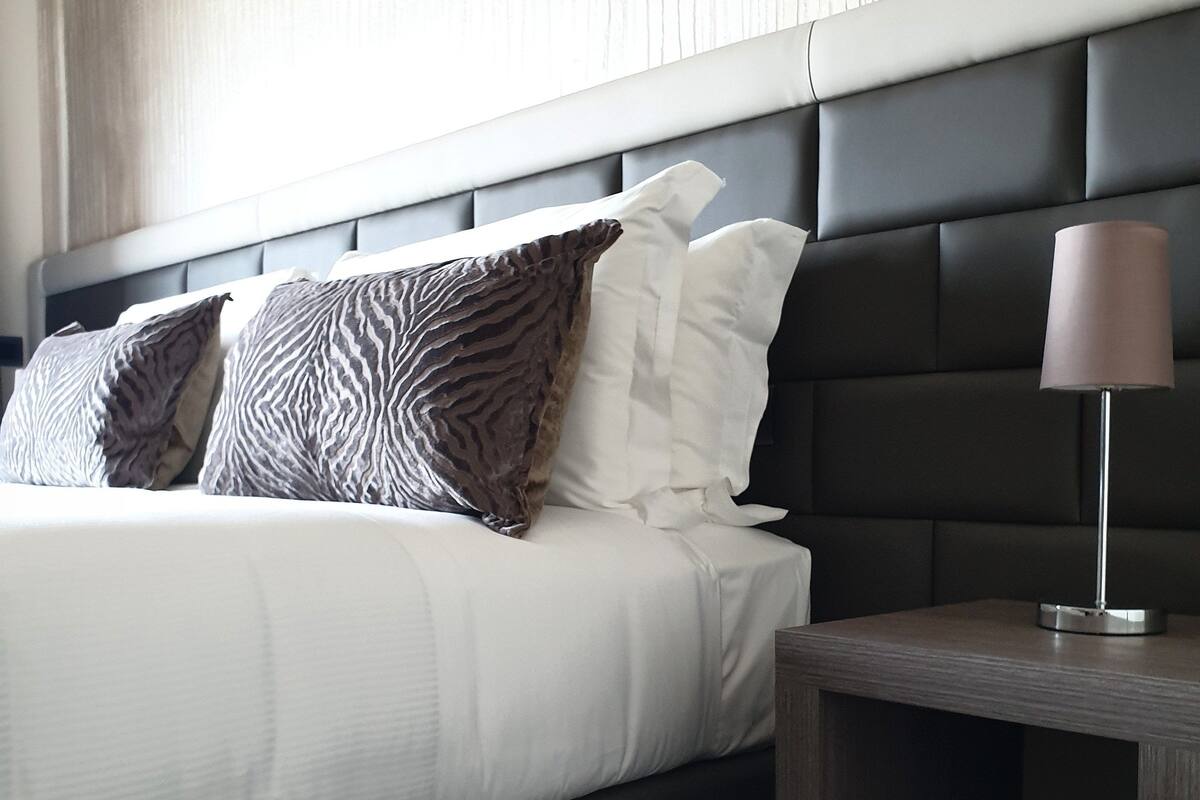 Una habitación de hotel moderna con una cama grande, cabecera oscura y una mesita de noche con lámpara.