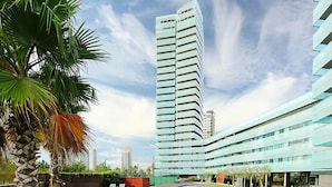 A modern hotel with a swimming pool, palm trees, and a tall building in the background. - Diagonal Mar by Interhome (Barcelona)