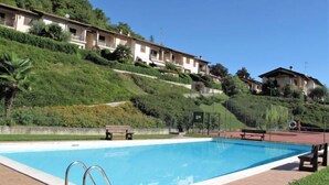 A swimming pool with a metal ladder, surrounded by a brick deck. There are benches and a grassy area nearby. - La Rocca by Interhome (Castelveccana)