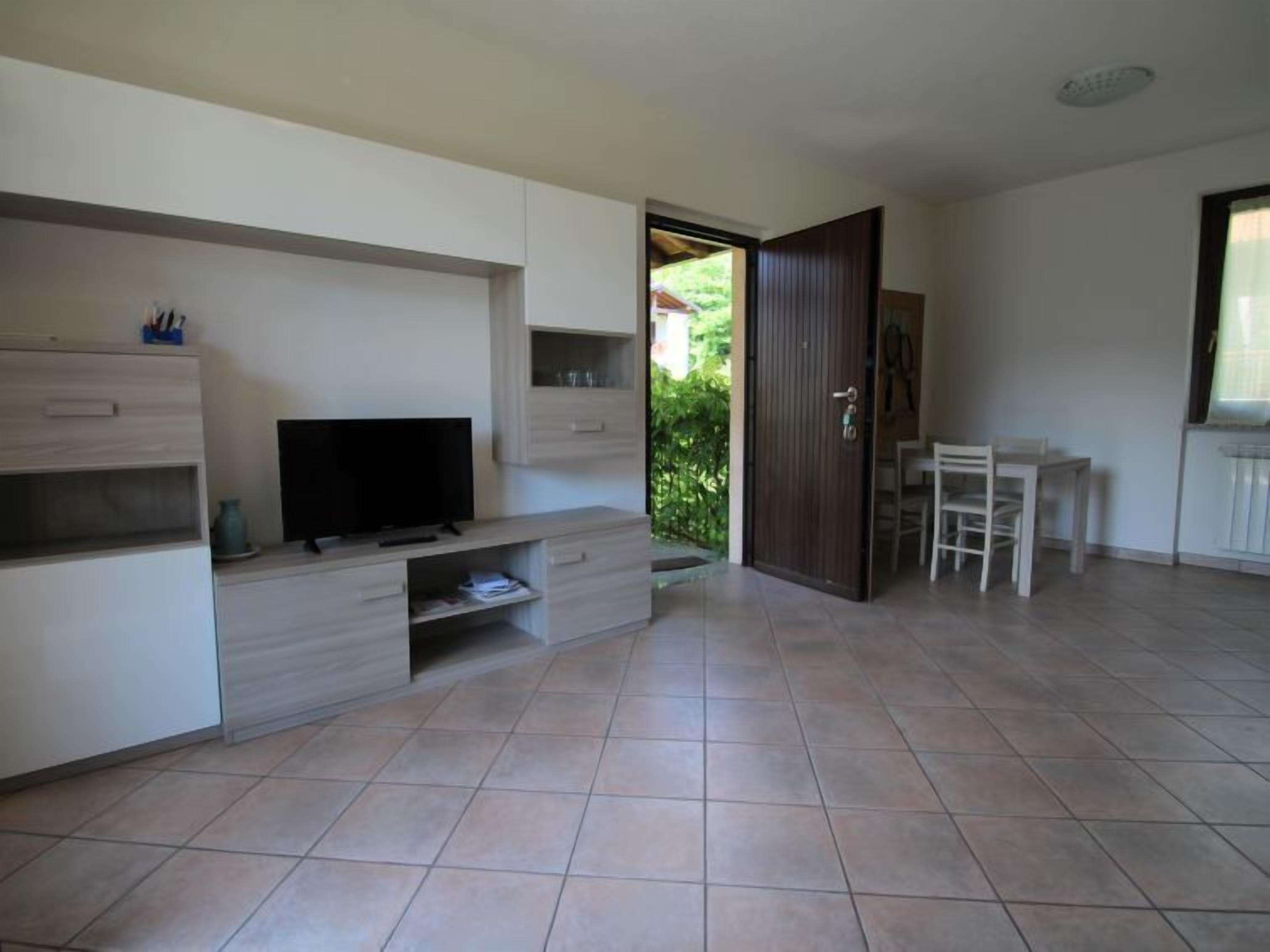 A modern living room with a flat-screen TV, built-in storage, a dining area with a table and chairs, and a view of greenery outside.
