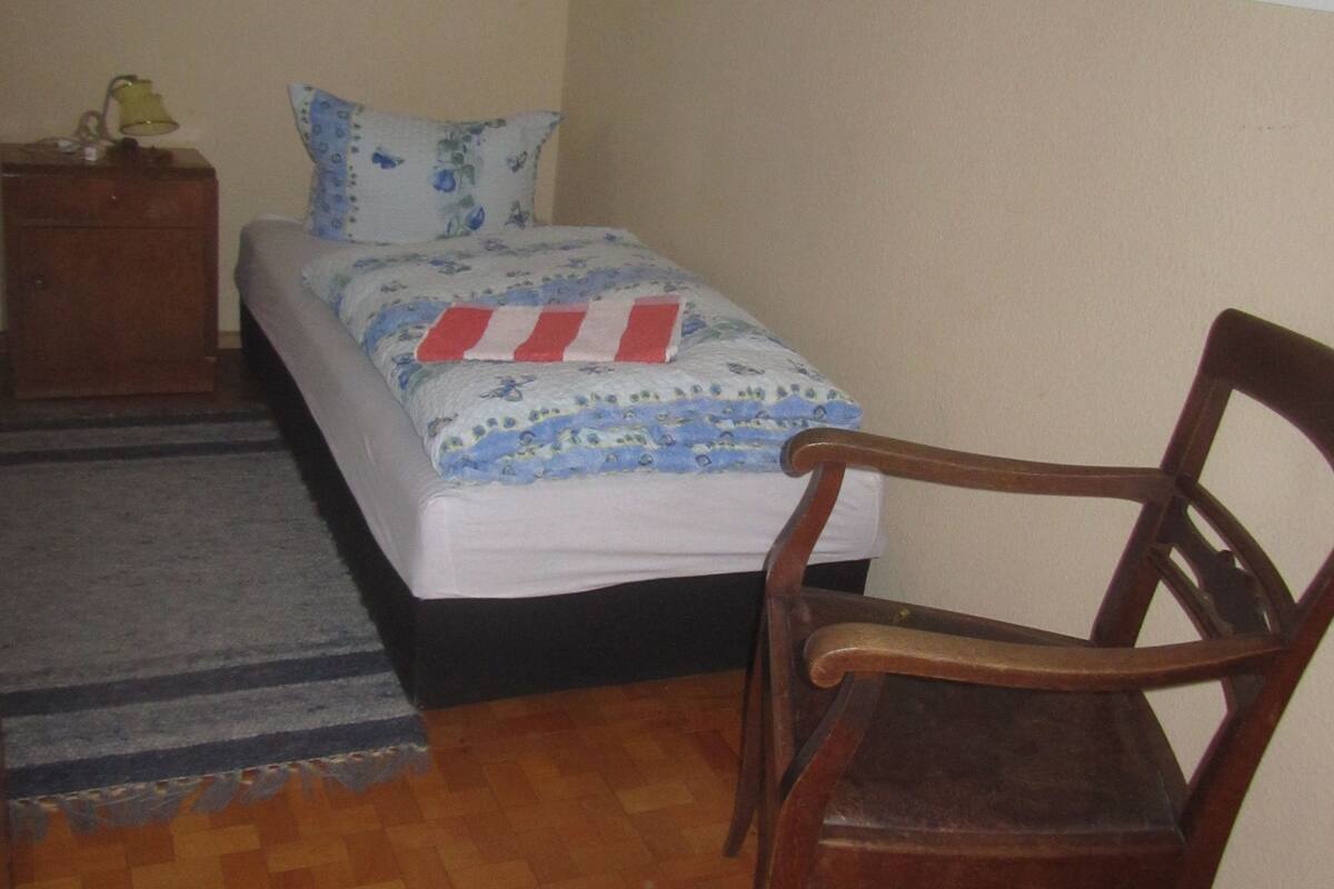 Ein Schlafzimmer mit einem Bett, einem Nachttisch aus Holz, einem Sessel, einem gemusterten Teppich und Holzdielen.