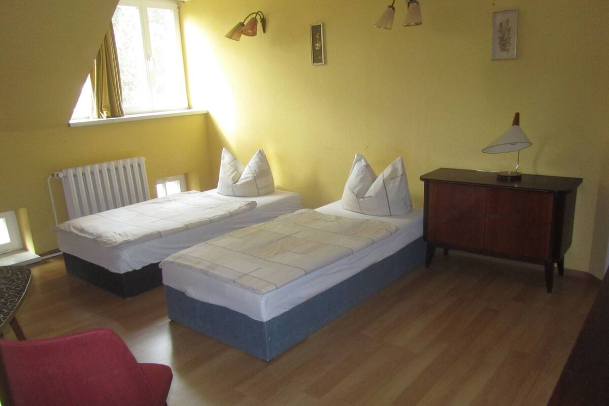 Ein Zimmer mit zwei Einzelbetten, Holzboden, einem roten Stuhl, einem Holzschrank und einem Fenster mit Vorhängen.