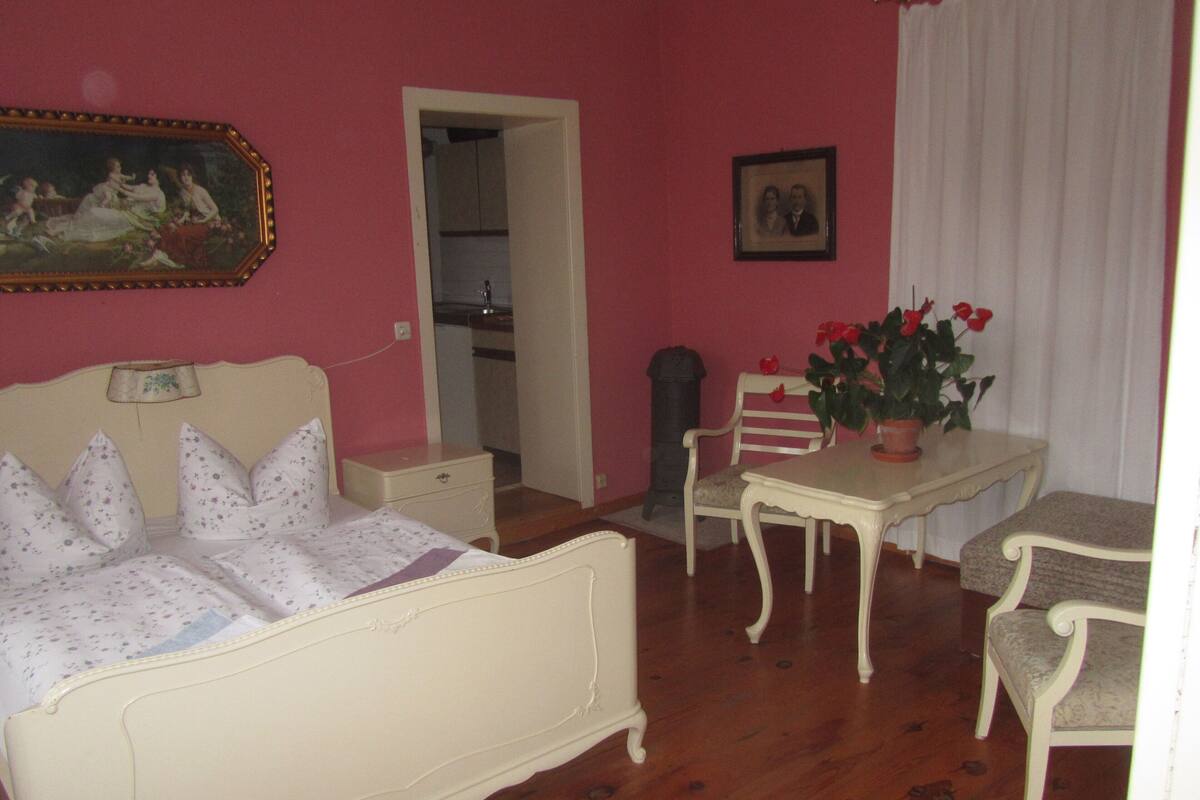 Ein Zimmer mit einem Bett, einem Tisch, einem Sessel, einer Zimmerpflanze und einem Bild an der Wand.