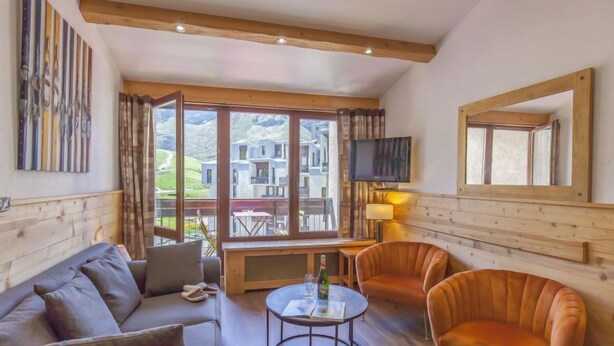 Ein modernes Wohnzimmer mit einer Couch, Sesseln, einem Couchtisch und einem Fernseher. Große Fenster bieten Blick auf Gebäude und Grünflächen.