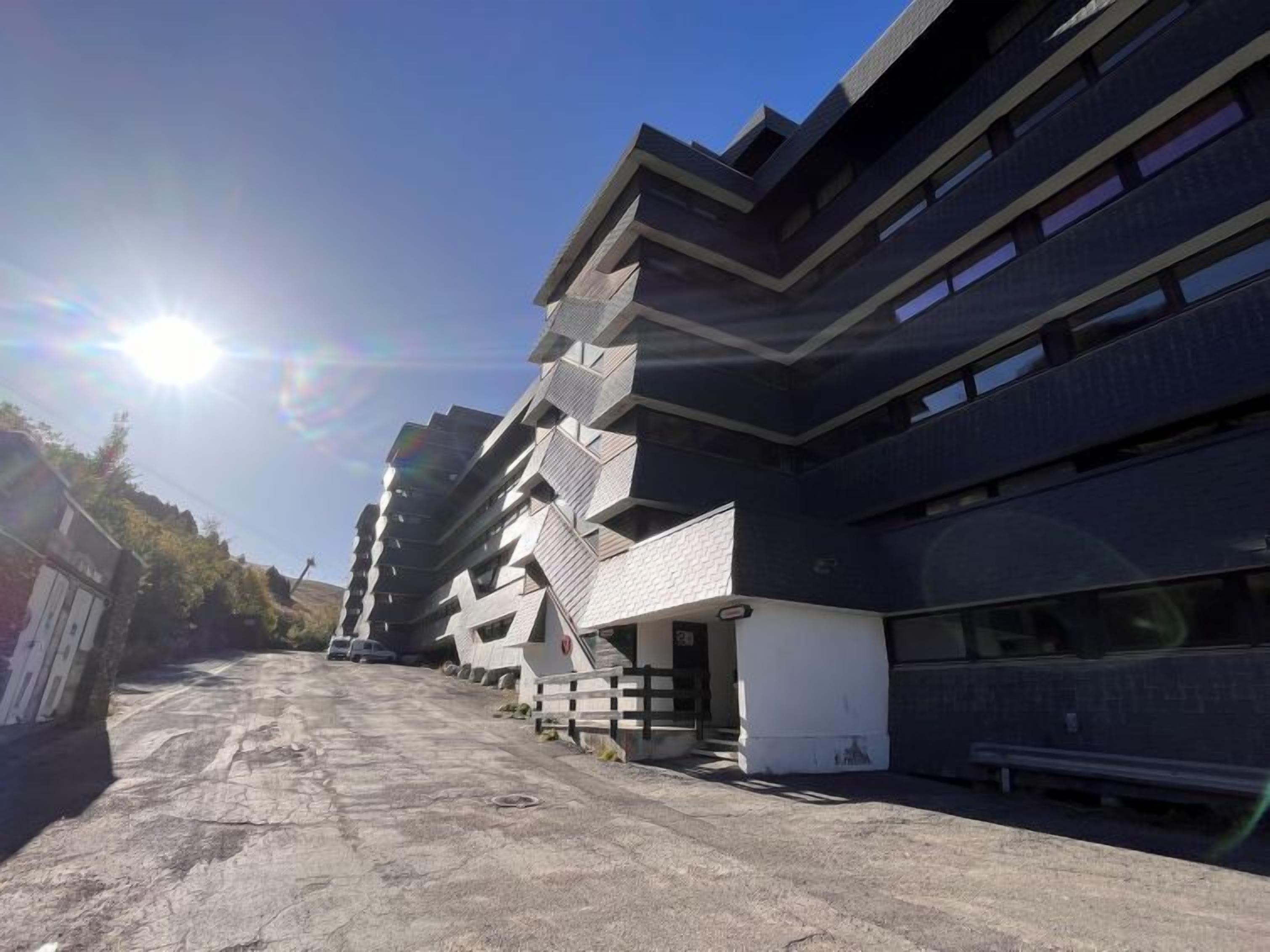 Un edificio moderno de varios pisos con un diseño angular distintivo, que cuenta con balcones y un estacionamiento al frente.