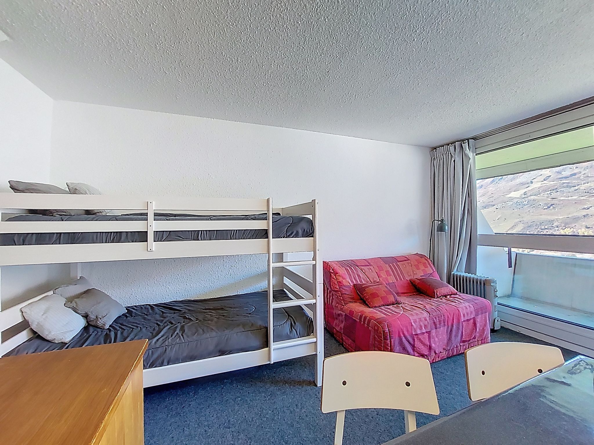Een slaapkamer met een stapelbed, een rode deken en kussens, een houten bureau en een raam met uitzicht op een landschap.