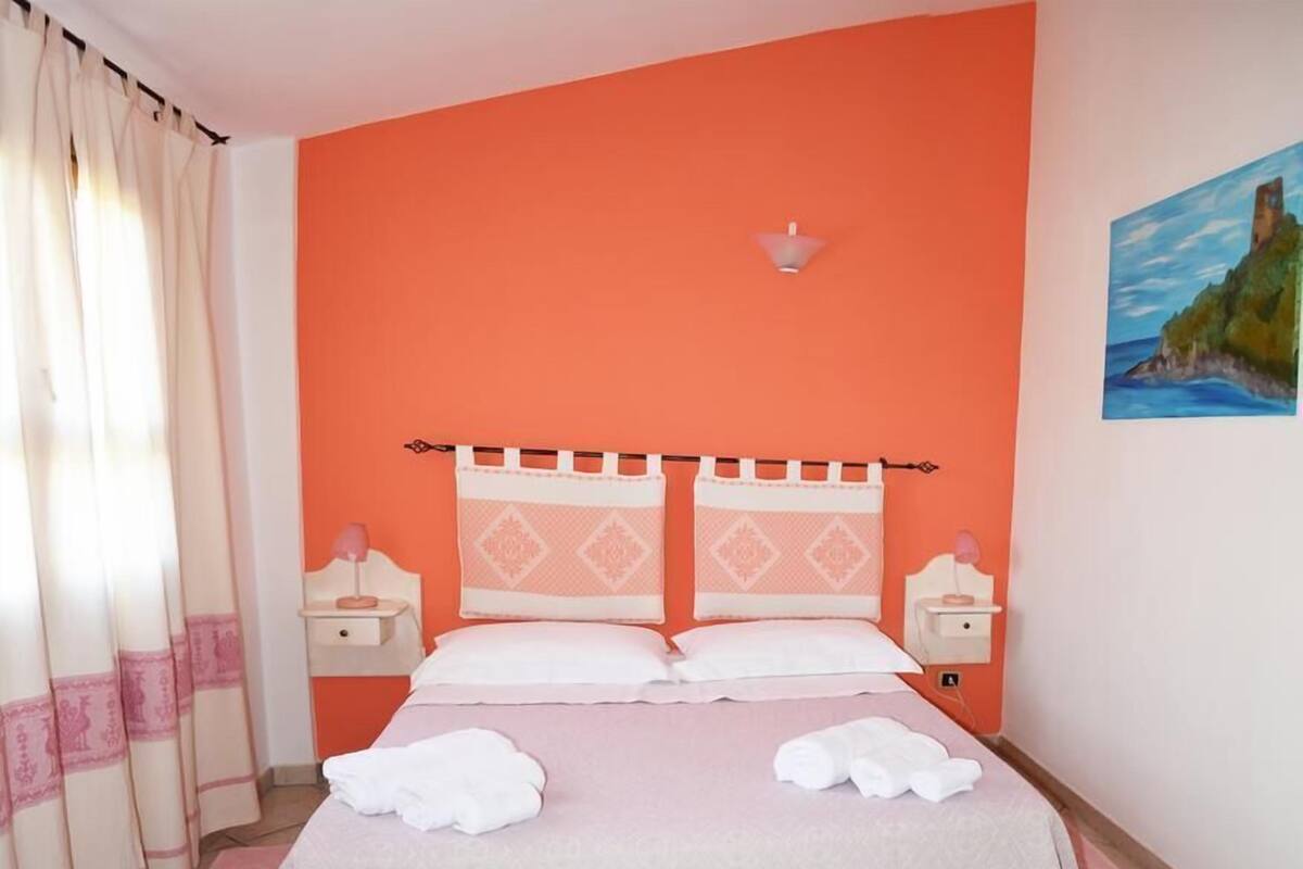 Une chambre avec un mur orange, un lit recouvert de draps blancs, deux tables de chevet avec des lampes et un tableau accroché au mur.