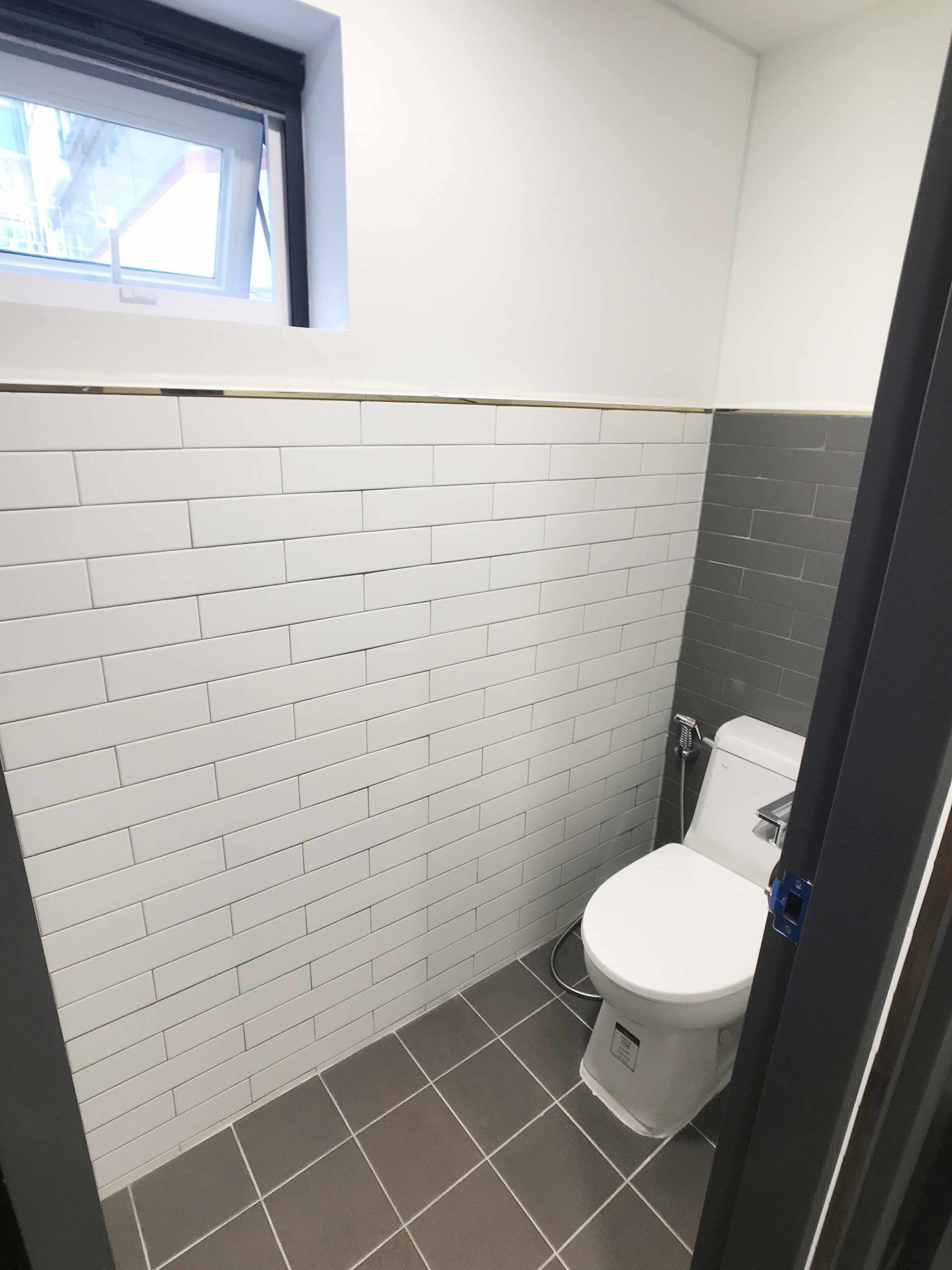 Een badkamer met wandtegels in wit en grijs, een wit toilet en een raam boven het toilet.