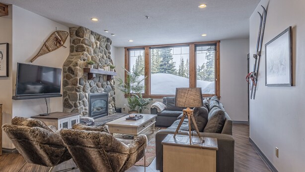 Uma sala de estar moderna com lareira de pedra, televisão de tela plana e uma janela grande com vista para uma paisagem nevada.
