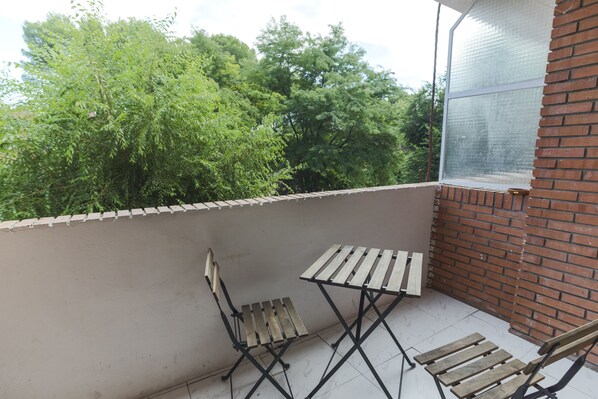 A balcony with a view of trees, a brick wall, and a window with a screen.