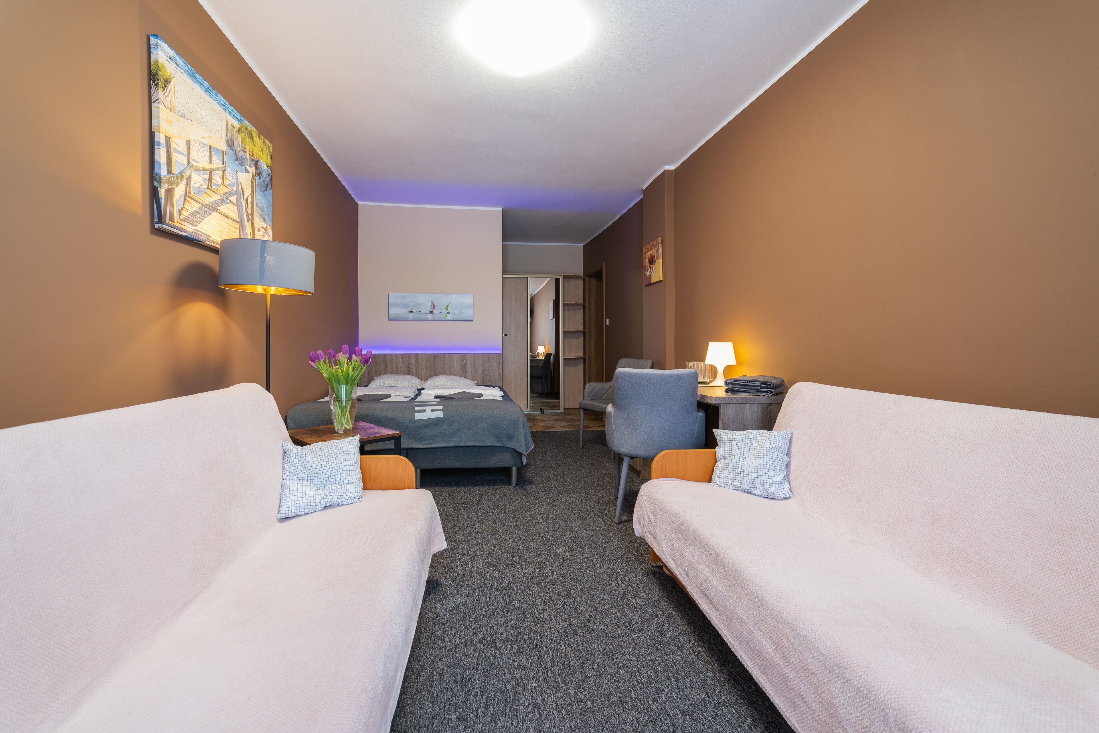 A hotel room with two single beds, a desk with a lamp, a chair, a small table with a vase of flowers, and a painting on the wall.