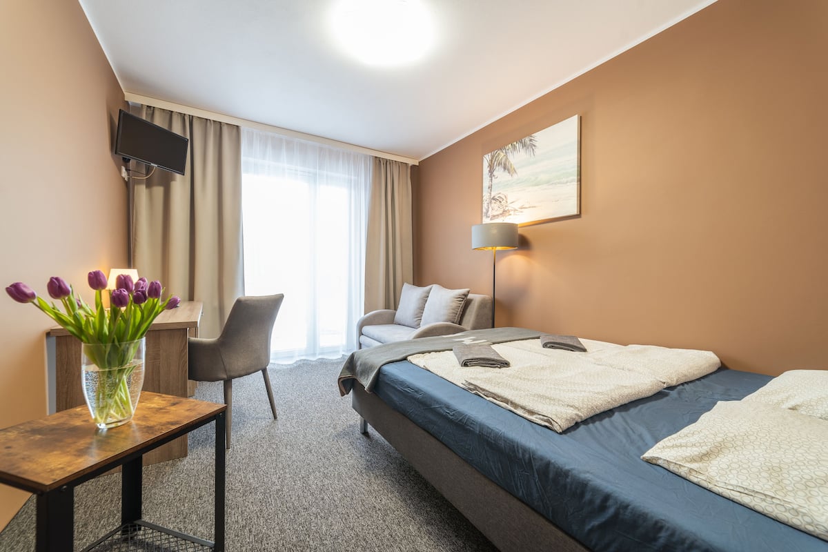 A hotel room with a bed, a desk, a chair, a TV mounted on the wall, a lamp, and a painting on the wall.