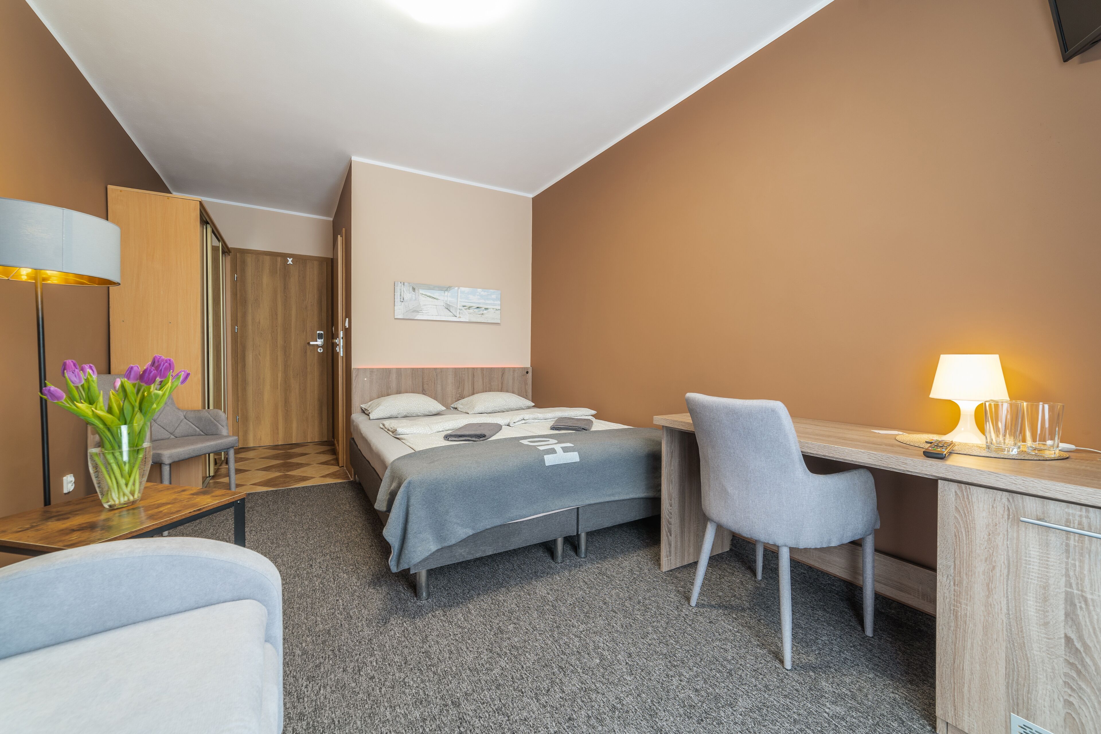 A hotel room with a bed, desk, chair, lamp, and a vase of flowers.