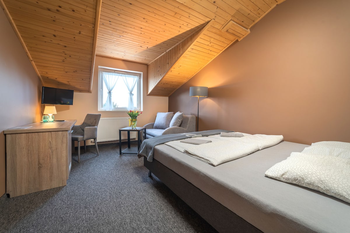 A bedroom with a wooden ceiling, a bed with white linens, a desk with a lamp, a chair, and a TV mounted on the wall.
