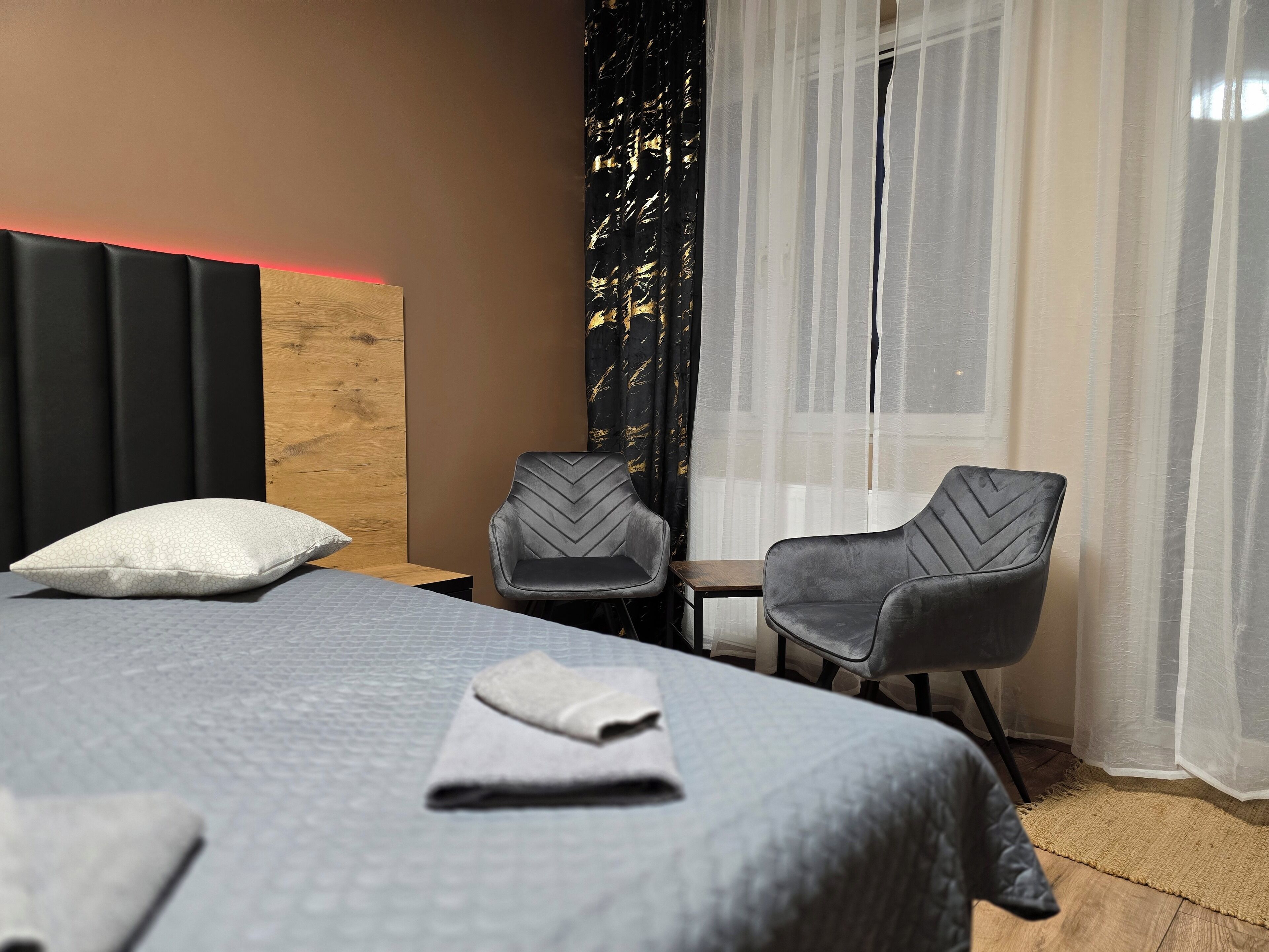 Een hotelkamer met een bed, twee stoelen, een houten hoofdbord en dunne gordijnen.