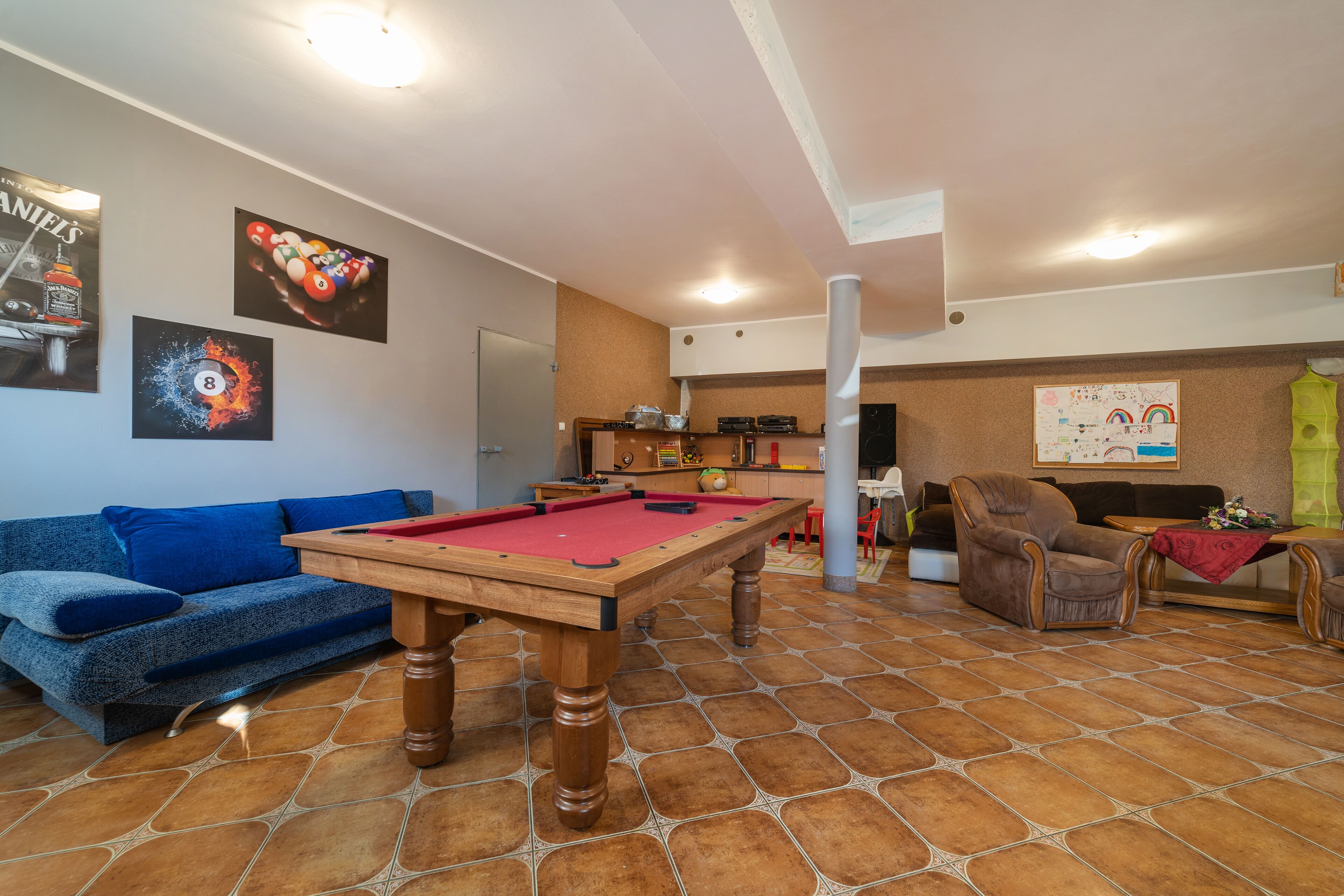 A living room with a pool table, a blue sofa, a brown armchair, and a kitchen area in the background.