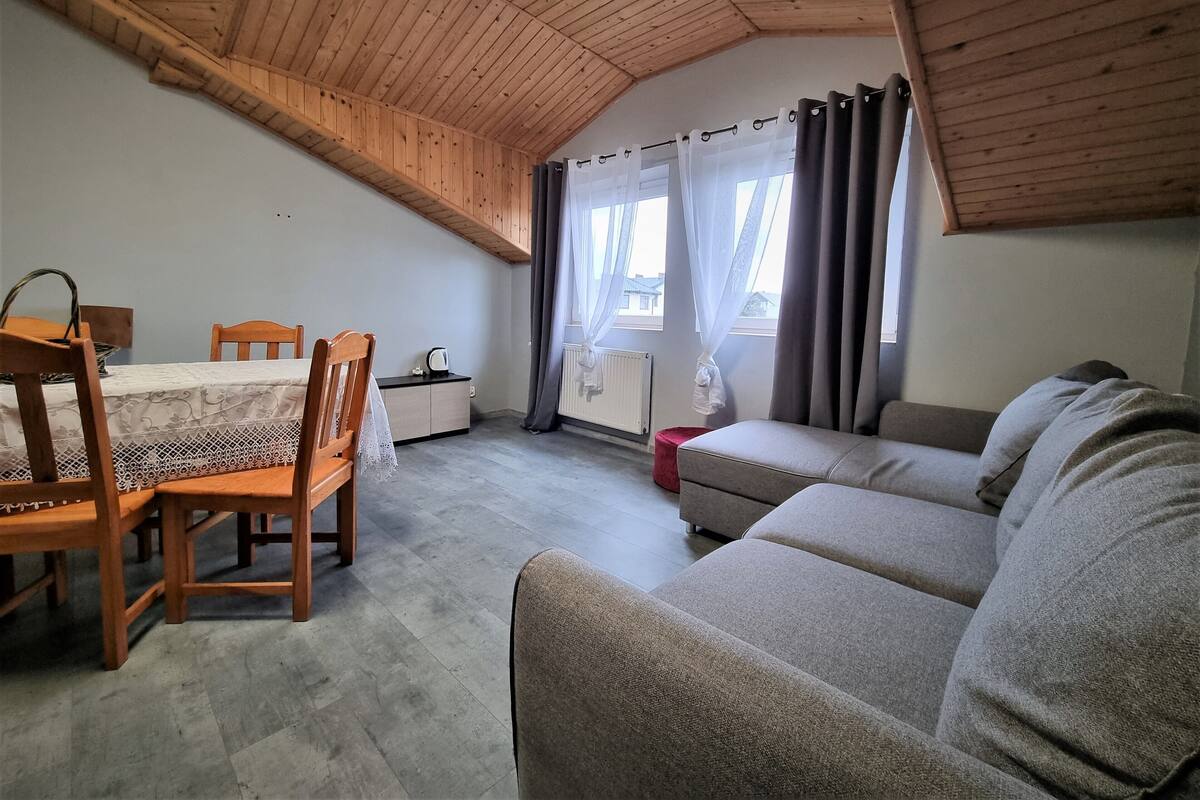 A cozy attic room with a wooden ceiling, a dining table with chairs, a sofa, and a small cabinet.