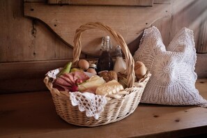 A wicker basket filled with various food items and a bottle, placed on a wooden bench with a crocheted pillow beside it. - Pension Aichner (Terento)
