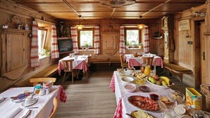Un coin repas rustique aux murs et au plafond en bois, avec plusieurs tables dressées pour le petit-déjeuner, où l'on peut trouver une variété de plats et de boissons.