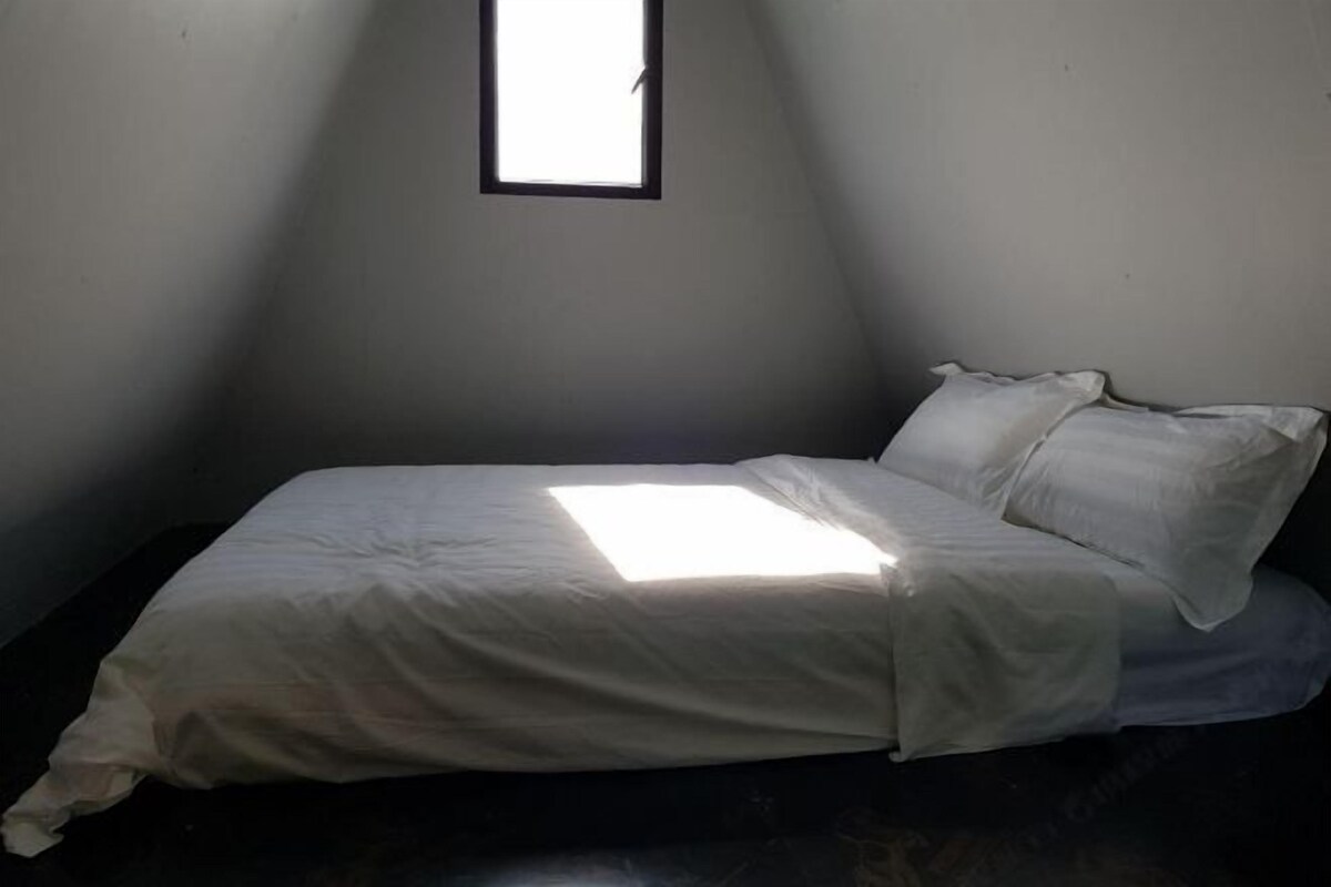 Ein Einzelbett in einem Raum mit schrägverlaufender Decke, kleinem Fenster und schlichten Wänden.