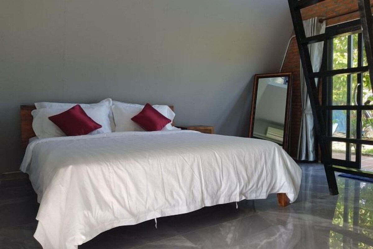 Quarto com cama, duas almofadas vermelhas, cabeceira de madeira, um espelho grande e vista para o verde lá fora.